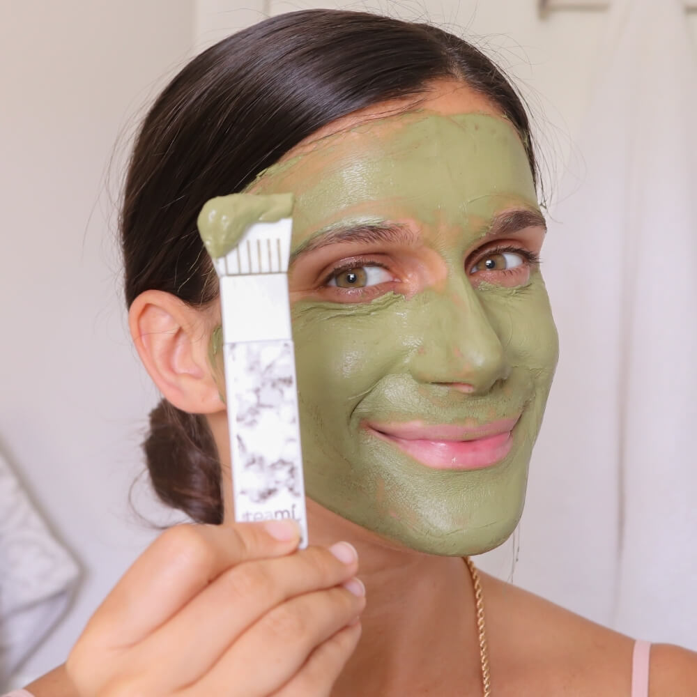 Woman with detox mask on face holding Teami face mask applicator brush