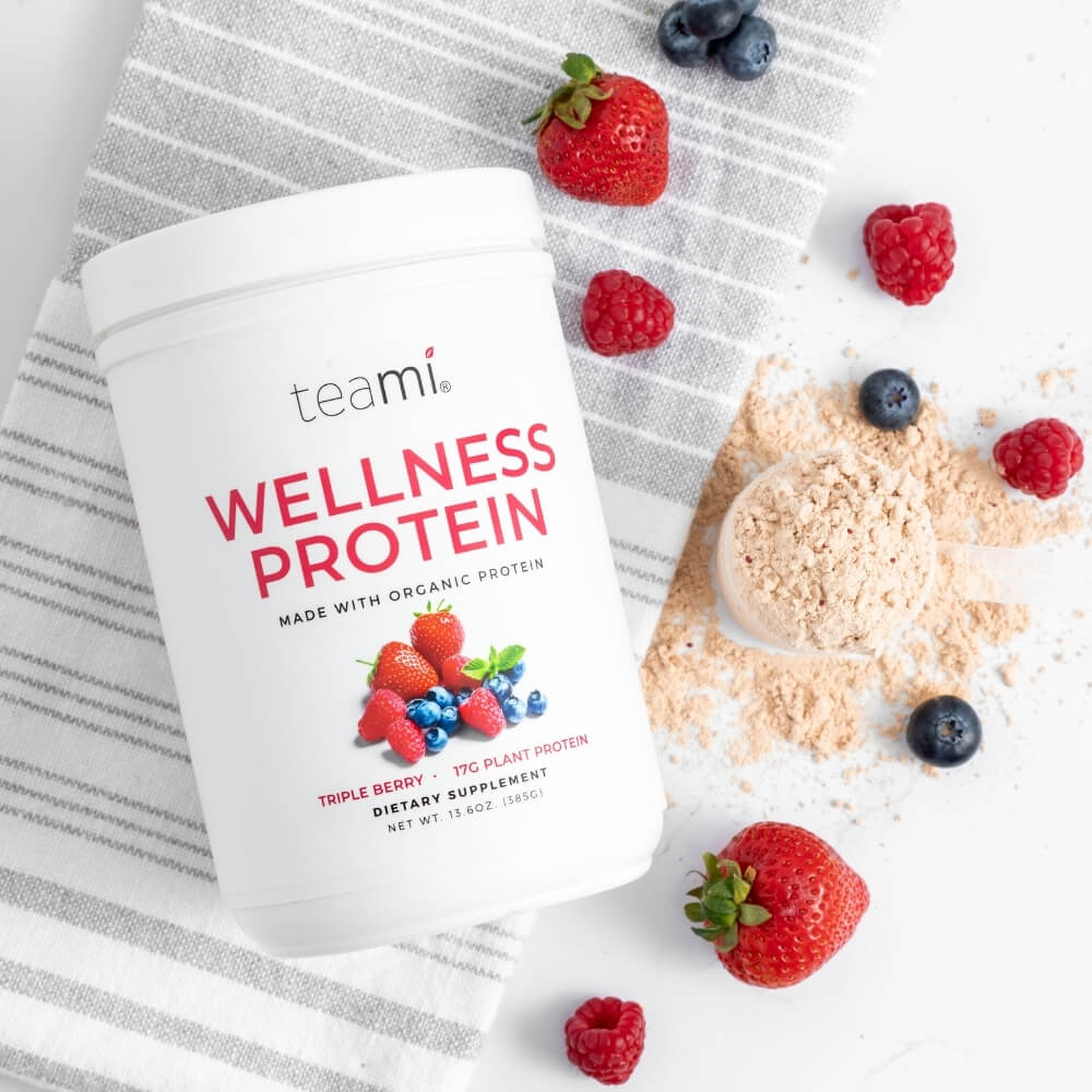Tub of Teami wellness protein triple berry flavour lying on kitchen surface with fruit and scoop of powder
