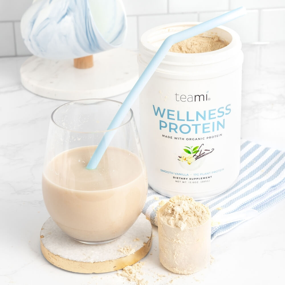 glass of protein shake with straw next to tub of teami wellness protein smooth vanilla flavour